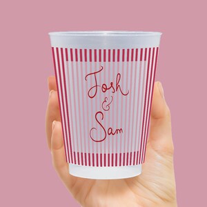 Op de afbeelding: Een witte beker met rode en roze verticale strepen, vastgehouden in een hand. De beker heeft de namen "Josh & Sam" in een sierlijk lettertype, perfect voor een bruiloft of feest. De beker is gemaakt van een doorschijnend materiaal.
