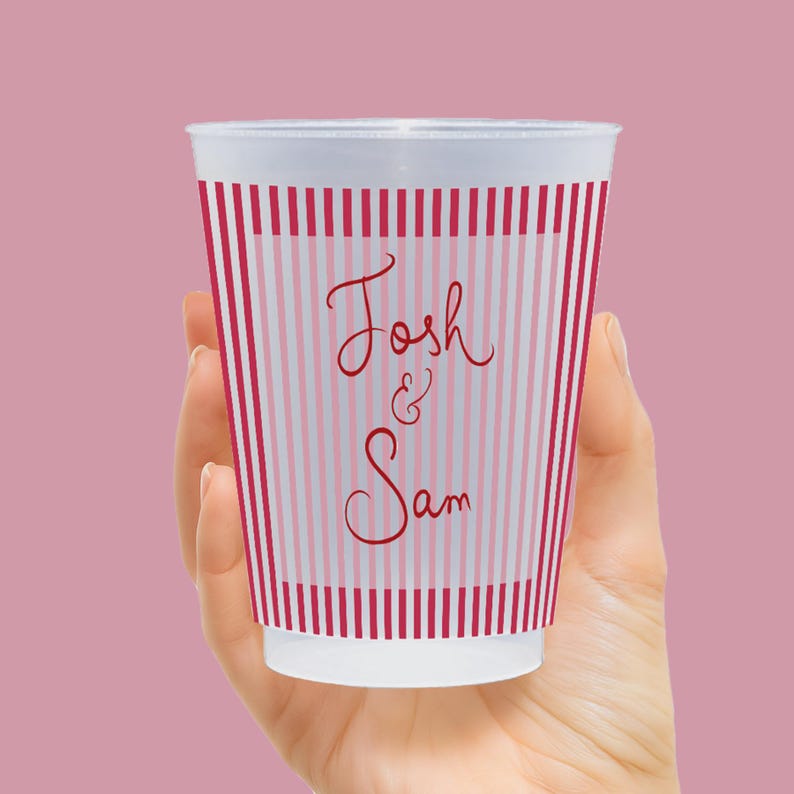 Op de afbeelding: Een witte beker met rode en roze verticale strepen, vastgehouden in een hand. De beker heeft de namen "Josh & Sam" in een sierlijk lettertype, perfect voor een bruiloft of feest. De beker is gemaakt van een doorschijnend materiaal.