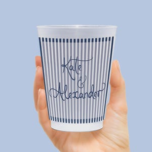 Op de afbeelding: Een witte plastic beker met een marineblauw gestreept patroon en de namen "Kate & Alexander" in cursief schrift. De beker wordt in een hand gehouden tegen een lichtblauwe achtergrond.