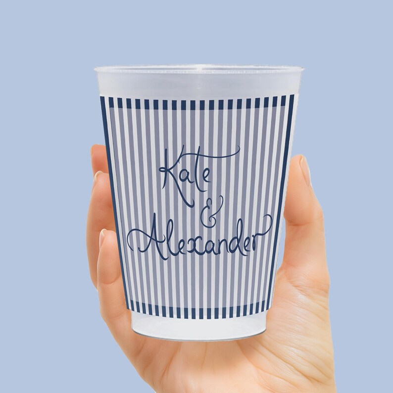 Op de afbeelding: Een witte plastic beker met een marineblauw gestreept patroon en de namen "Kate & Alexander" in cursief schrift. De beker wordt in een hand gehouden tegen een lichtblauwe achtergrond.