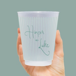 Op de afbeelding: Een doorzichtige plastic beker met verticale lichtblauwe strepen. De beker heeft de namen "Harper + Luke" in een turquoise scriptlettertype. De beker wordt in een hand gehouden tegen een effen teal achtergrond.