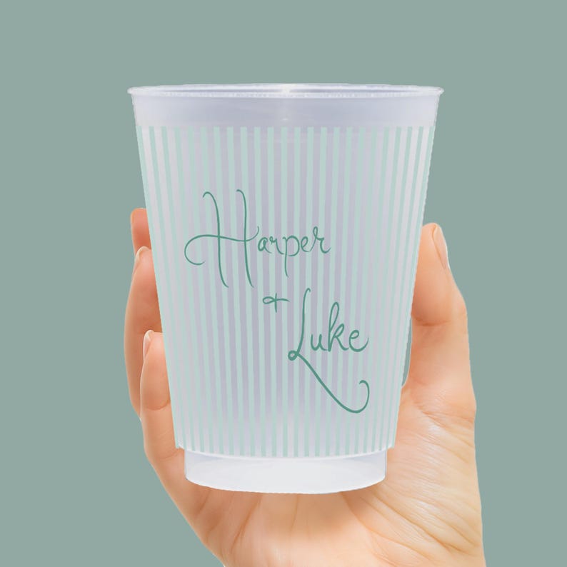 Op de afbeelding: Een doorzichtige plastic beker met verticale lichtblauwe strepen. De beker heeft de namen "Harper + Luke" in een turquoise scriptlettertype. De beker wordt in een hand gehouden tegen een effen teal achtergrond.