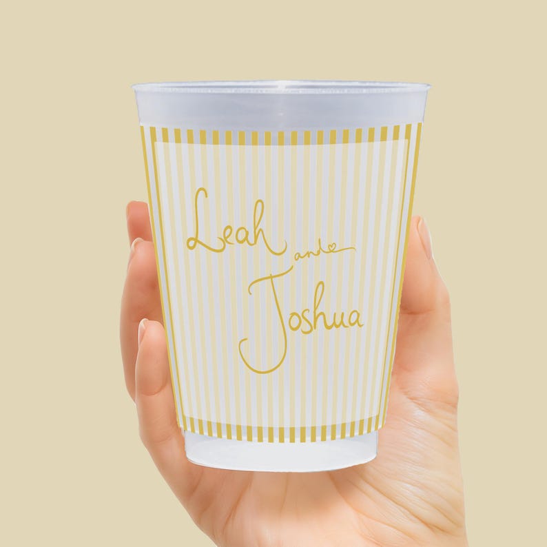 Op de afbeelding: Een wit en goud gestreepte wegwerpbeker met de namen "Leah and Joshua" in gouden script. De beker wordt in een hand gehouden en heeft een witte rand en basis. De beker is ontworpen voor een speciale gelegenheid.