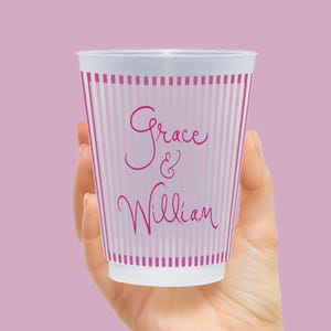 Op de afbeelding: Een witte wegwerpbeker met roze verticale strepen en de namen "Grace & William" in cursief schrift. De beker wordt in een hand gehouden tegen een effen roze achtergrond. De beker heeft een eenvoudig, elegant ontwerp.