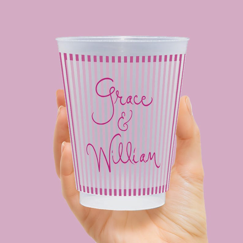 Op de afbeelding: Een witte wegwerpbeker met roze verticale strepen en de namen "Grace & William" in cursief schrift. De beker wordt in een hand gehouden tegen een effen roze achtergrond. De beker heeft een eenvoudig, elegant ontwerp.
