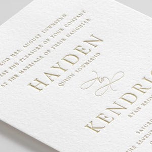 May include: Close-up of a white invitation with gold embossed text. The text includes names and details for a wedding, such as "Hayden" and "Kendri". The paper has a textured finish, and the overall design is elegant and formal.