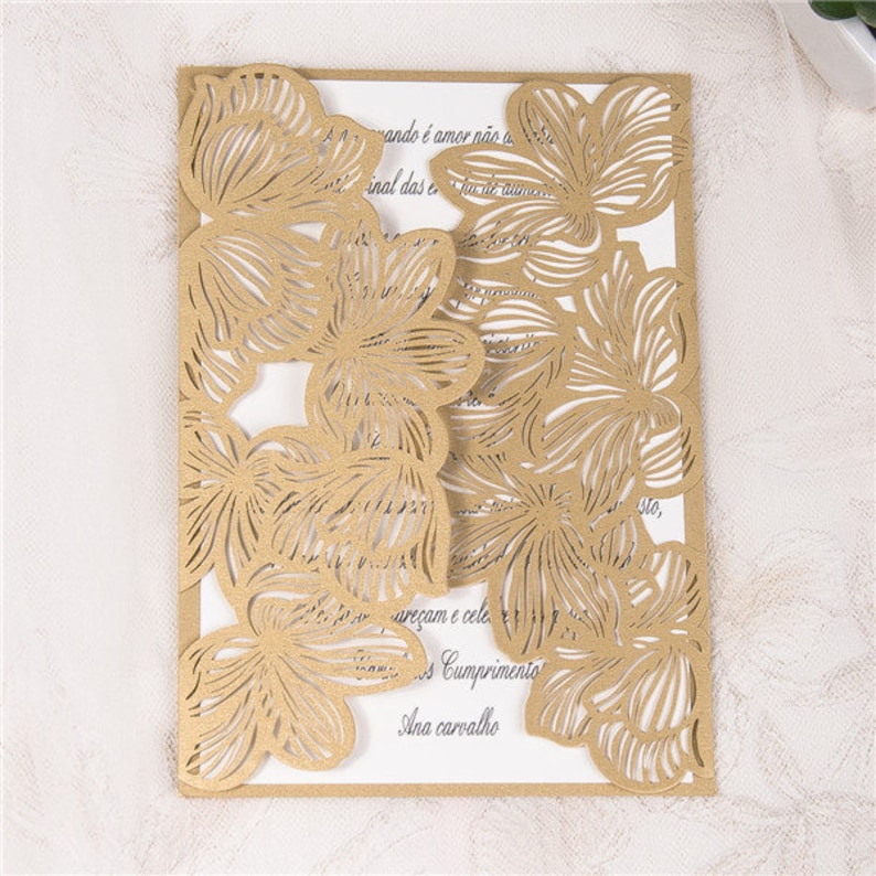 May include: Gold laser cut wedding invitation with a floral design. The invitation features a delicate pattern of flowers and leaves, creating a romantic and elegant look. The text on the invitation reads "ando &eacute; amor n&atilde;o a", "inal das e de au", "are&ccedil;am e cele", "Cumprimento", and "Ana carvalho".
