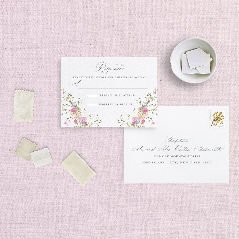 May include: A white RSVP card with floral border and text that reads "Response" and "Kindly reply before the thirteenth of May". The card has two lines for guests to indicate if they will attend or regretfully decline. The card is next to a white envelope with a handwritten address that reads "The future Mr and Mrs Collin Starworth 1229 Oak Mountain Drive Long Island City, New York 11101".