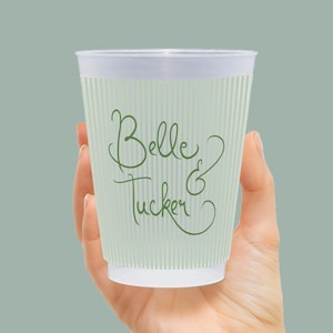 Op de afbeelding: Een doorzichtige plastic beker met verticale groene strepen en de woorden "Belle & Tucker" in groen schrift. De beker wordt in een hand gehouden tegen een effen groene achtergrond. De beker lijkt leeg te zijn.