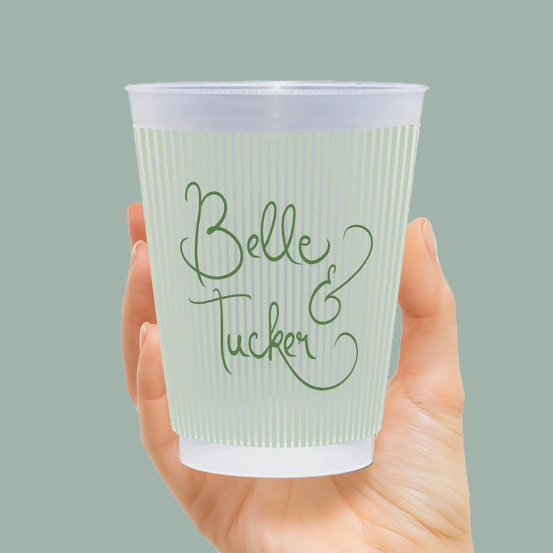 Op de afbeelding: Een doorzichtige plastic beker met verticale groene strepen en de woorden "Belle & Tucker" in groen schrift. De beker wordt in een hand gehouden tegen een effen groene achtergrond. De beker lijkt leeg te zijn.