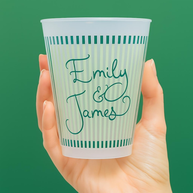 Op de afbeelding: Een doorzichtige plastic beker met een groen en wit gestreept ontwerp. De beker heeft de namen "Emily & James" in een turquoise scriptlettertype. De beker wordt in een hand gehouden tegen een groene achtergrond.