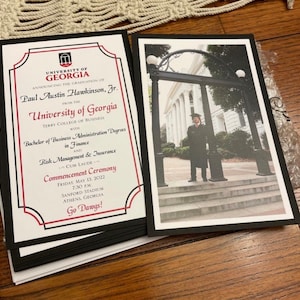 May include: A stack of graduation announcements from the University of Georgia, featuring a photo of a graduate and text announcing the commencement ceremony. The announcements have a black border and include the text "Go Dawgs!"