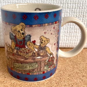 May include: A ceramic coffee mug with a blue and white border and a scene of two teddy bears baking a pie. The text "Sweetie Pies" is on the front of the mug.