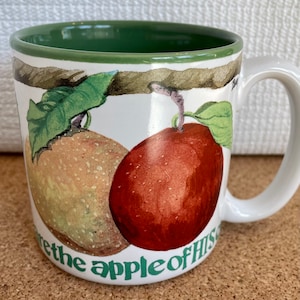 May include: White ceramic mug with a green rim. The mug features a watercolor illustration of two apples on a branch with green leaves. The text "You are the apple of my eye" is written in green on the bottom of the mug.