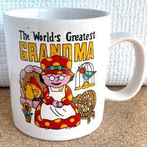 May include: White ceramic mug with a colorful illustration of a grandmother figure with a spinning wheel, a bird in a cage, and a fireplace. The text "The World's Greatest Grandma" is printed in black on the mug.