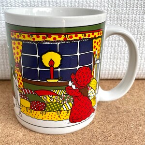May include: White ceramic mug with a colorful illustration of a girl in a red dress praying in bed with a lit candle in the window behind her.