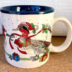 May include: White ceramic Christmas mug with a blue rim. The mug features a festive design of a duck wearing a Santa hat and holding a red ribbon. The duck is surrounded by holly, presents, and snowflakes.