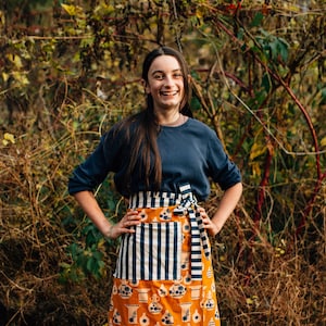 Może przedstawiać: Pomarańczowy fartuch z biało-granatowym paskiem na górze i wzorzystym dołem z wazonami i kwiatowymi wzorami. Fartuch jest zawiązany w talii. Osoba ma na sobie granatową koszulę z długim rękawem.