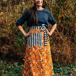 May include: A long wrap skirt featuring an orange base with a repeating dark blue pattern of various pottery shapes. The skirt includes a wide waistband and a large patch pocket, both decorated with a dark blue and white vertical stripe design.