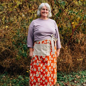 May include: An orange apron with a white and blue vase pattern, paired with a striped pocket. The apron is tied at the waist and worn over a long-sleeved lilac shirt and dark blue trousers. The person is wearing brown shoes.