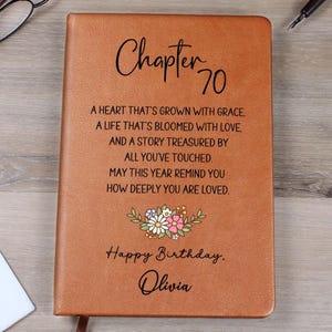 May include: A brown leather-bound journal with the text "Chapter 70" and a personalized birthday message. The cover includes a floral design and the name "Olivia". The journal is open, revealing the lined pages.