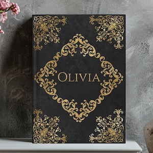 May include: A black journal with gold filigree corner and border designs. The word "OLIVIA" is in gold script within a gold diamond-shaped frame on the cover. The journal is displayed on a white surface.
