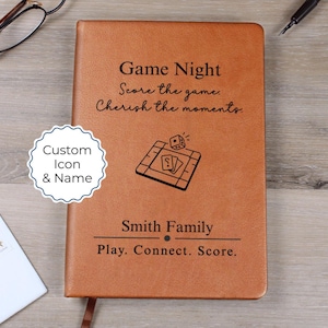 Puede incluir: Un diario de cuero marrón con las palabras "Game Night" y "Smith Family" grabadas en la portada. El diario presenta una ilustración de un juego de mesa y el texto "Play. Connect. Score."