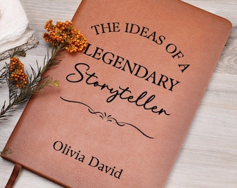 May include: A brown leather journal with the text "THE IDEAS OF A LEGENDARY Storyteller" and the name "Olivia David" printed in black. A sprig of orange flowers rests on the journal. The journal has a ribbon bookmark.