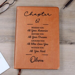 May include: A brown leather journal with the text "Chapter 67" and inspirational quotes. The journal is personalized with the name "Olivia" at the bottom. A bookmark is visible. The journal is on a wooden surface.