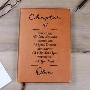 May include: A brown leather journal with the text "Chapter 47" and inspirational phrases. The journal includes the name "Olivia" at the bottom. A pen and glasses are visible in the background.