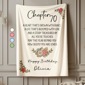 May include: A cream-colored blanket with the text "Chapter 70" and a birthday message. Floral designs in pink, white, and green adorn the corners and center. The message includes the name "Olivia".