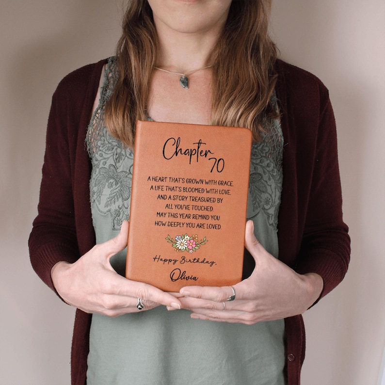 May include: A tan-colored book with the words "Chapter 70" and a poem printed on the cover. The book also includes the text "Happy Birthday, Olivia" and a floral design. The book is held by a person.