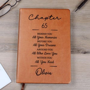 Pode incluir: Um diário de couro castanho com o texto "Capítulo 65" e frases inspiradoras. O diário inclui o nome "Olivia" na parte inferior. Uma fita de marcador é visível.