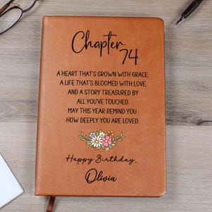 Puede incluir: Un diario de cuero marrón con las palabras "Chapter 74" y un diseño floral. El diario incluye el texto "Happy Birthday, Olivia" y un mensaje sincero sobre el amor y la gracia.