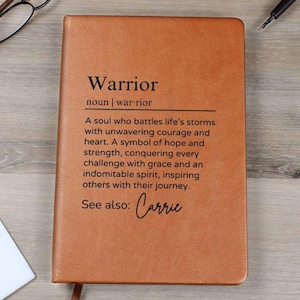 Puede incluir: Un diario de cuero marrón con la palabra "Warrior" definida como "Un alma que lucha contra las tormentas de la vida con un coraje inquebrantable y un corazón fuerte." El texto "Ver también: Carrie" está en la parte inferior del diario.