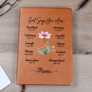 May include: A brown leather journal with the words "God Says You Are" and inspirational phrases like "Beautiful" and "Amazing." A floral design with a pink flower and green leaves is in the center. The name "Maria" is at the bottom.