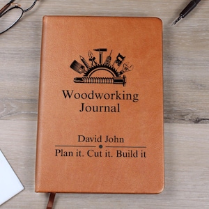 Puede incluir: Un diario de carpintería encuadernado en cuero marrón con el título "Woodworking Journal" y el nombre "David John" impresos en la portada. La portada también presenta un gráfico de herramientas de carpintería. El texto "Plan it. Cut it. Build it" está impreso debajo.