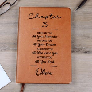 May include: A brown leather-bound journal with the text "Chapter 25" and inspirational phrases. The name "Olivia" is inscribed at the bottom. A bookmark is visible.