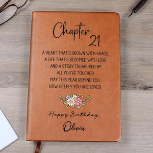 May include: A tan leather-like journal featuring the black printed text Chapter 21, a poem about love and grace, a small colourful floral illustration, the phrase Happy Birthday, and the name Olivia centred on the front cover.