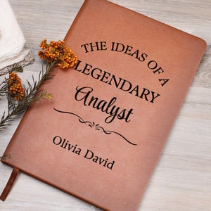 Może przedstawiać: Brązowy skórzany notatnik z czarnym napisem "THE IDEAS OF A LEGENDARY Analyst Olivia David". Na notatniku leży ozdobna gałązka pomarańczowych kwiatów i zieleni. Notatnik ma wstążkę do zaznaczania stron.