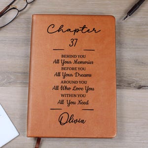 May include: A brown leather journal with the text "Chapter 37" and inspirational phrases. The name "Olivia" is at the bottom. The journal has a ribbon bookmark.