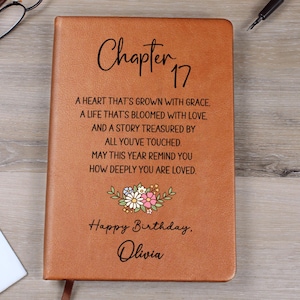 May include: A brown leather journal with the words "Chapter 17" and a personalized birthday message. The cover features a floral design and the name "Olivia". The journal is closed with a ribbon.