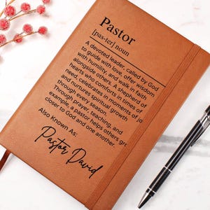 Puede incluir: Un diario de cuero marr&oacute;n con la palabra "Pastor" definida como un l&iacute;der devoto llamado por Dios para guiar con amor, ofrecer sabidur&iacute;a y caminar en la fe junto a otros. El diario tambi&eacute;n tiene el texto "Also Known As: Pastor David" escrito en cursiva.