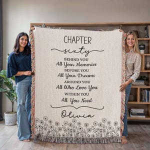 May include: Cream-colored woven throw blanket with black text that reads "Chapter Sixty" and a personalized name. The blanket has a floral border and colorful fringe.
