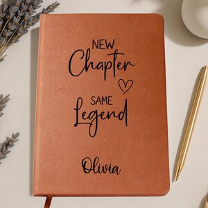 May include: A brown leather-bound journal with the words "New Chapter, Same Legend" and the name "Olivia" in black script. A gold pen rests beside the journal, and a sprig of lavender is in the background.