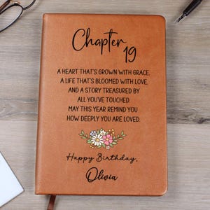 May include: A brown leather journal with the text "Chapter 19" and a birthday message. The journal includes the words "Happy Birthday, Olivia" and a floral design. A ribbon bookmark is visible.