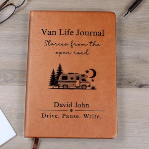 May include: A brown leather journal with the text "Van Life Journal" and "Stories from the open road." The cover features an illustration of a camper van, trees, a crescent moon, and stars. The name "David John" and the phrase "Drive. Pause. Write." are also printed on the cover.