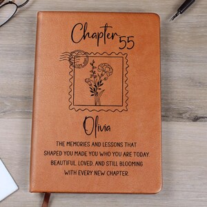 May include: A brown leather journal with the text "Chapter 55" and the name "Olivia" printed on the cover. The cover also features a floral design within a postage stamp frame and inspirational text.