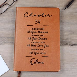 May include: A brown leather journal with the text "Chapter 54" and inspirational quotes. The name "Olivia" is also inscribed. The journal has a ribbon bookmark and is on a wooden surface.