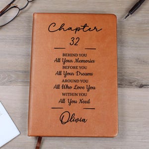 May include: A brown leather journal with the text "Chapter 32" and inspirational quotes. The journal is personalized with the name "Olivia" at the bottom. A pen and glasses are visible in the background.